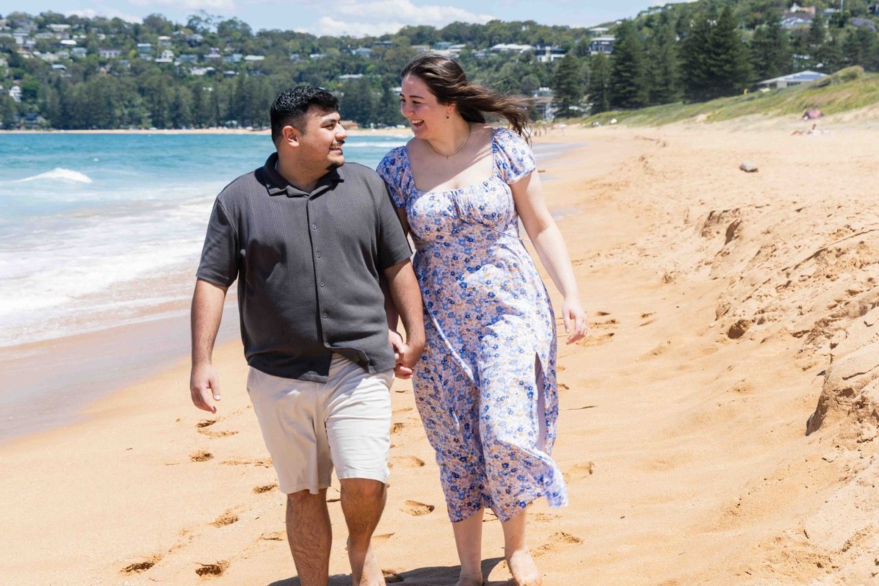 The couple smile at each other as they walk on the sand